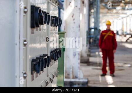 Electric switchers board. Focus on foreground. Worker in red work wear and yellow hard had blurred. Petrochemical Oil refinery plant. CNPC company. Stock Photo