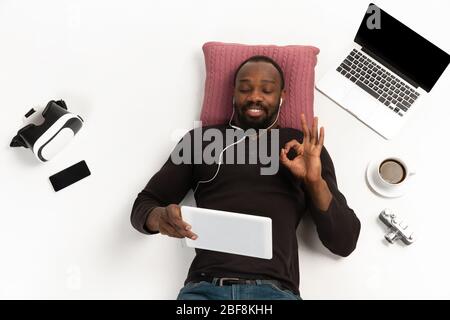 Young african-america man using devices, gadgets isolated on white studio background. Concept of modern technologies, gadgets, tech, emotions, advertising. Copyspace. Selfie, vlog, greeting education Stock Photo