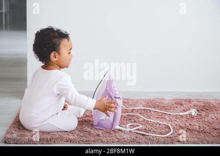 Little African-American baby playing with socket and iron at home. Child in danger Stock Photo