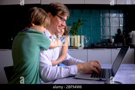 Remote home work during the quarantine period of the Covid 19 coronavirus, when the family is at home. A man works in the kitchen for a laptop. Father and children are smiling. Stock Photo