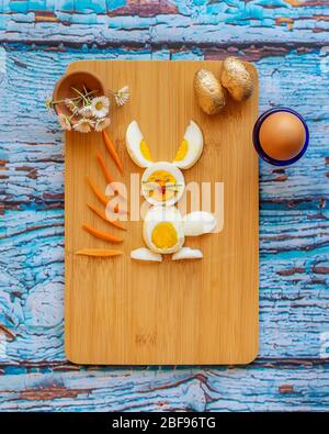 Beautiful bunny made with boiled eggs, carrots, rosemary and goji berry, paired with daisies and small Easter eggs, on a wooden cutting board and colo Stock Photo
