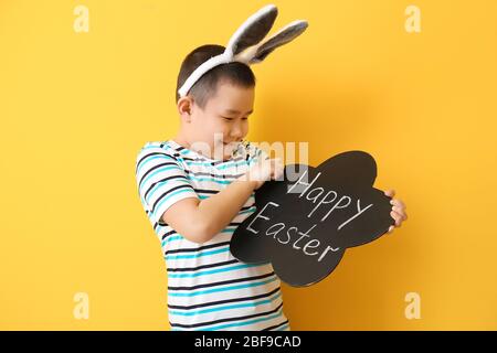 Little Asian boy holding chalkboard with text HAPPY EASTER on color background Stock Photo