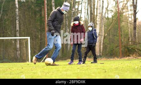 Family plays football on the playground during quarantine Covid-19. Father with children wearing medical face mask to protect from Corona Virus. Stock Photo