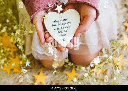 A girl or woman holding a heart with the quote when it rains look for rainbows, surrounded by gold stars. Coronavirus lockdown positive uplifting