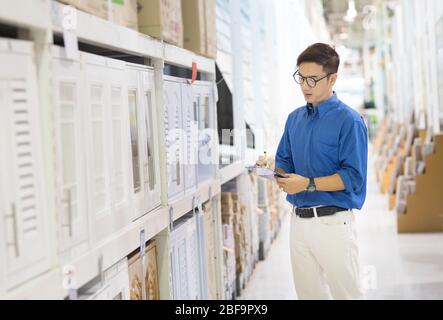 Asian manager man doing stocktaking of products management in cardboard box on shelves in warehouse using clipboard file and pen. Male professional as Stock Photo