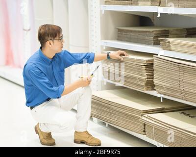 Asian manager man doing stocktaking of products management in cardboard box on shelves in warehouse using clipboard file and pen. Male professional as Stock Photo
