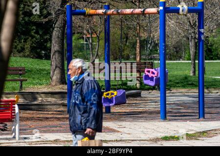 Senior man wearing protective face mask walking past empty deserted blue swings at colourful playground or play area. The public park playgrounds have been closed during the lockdown because of the Coronavirus Pandemic of Covid-19 spread as of April 2020 in Sofia, Bulgaria, Eastern Europe, Balkans, EU Stock Photo