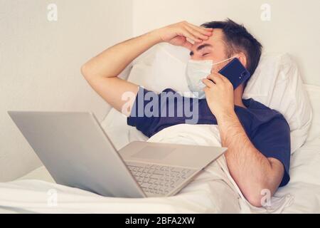 A patient in a hospital room with a headache tries to work at a computer. man sick coronavirus covid19 virus working at computer and wearing mask prot Stock Photo