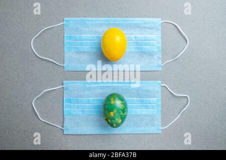 Yellow and green easter eggs lie on blue medical masks on a gray background. Flat lay. Easter holiday concept during quarantine coronavirus epidemic. Stock Photo