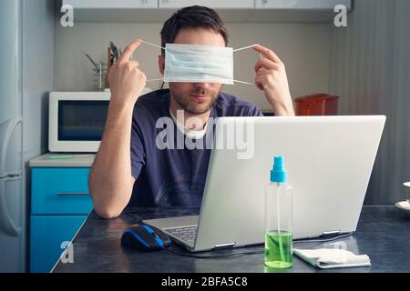 A man is sitting at a table and working on a laptop in home clothes against the background of the kitchen. Tired of remote work and quarantine, the gu Stock Photo