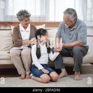 Asian senior Grandmother and happy asia elder grandfather sitting on sofa playing and enjoying leisure time with granddaughter in living in living at Stock Photo