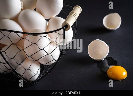 Chicken eggs in a metal basket on a black wooden table. Cracked egg directly on table with the yellow yolk and broken eggshell. Concept image for the Stock Photo