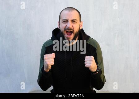A guy with a beard clenching his fists screams. A man looks at the camera clenching his fists and opening his mouth, an emotion of aggression, victory Stock Photo