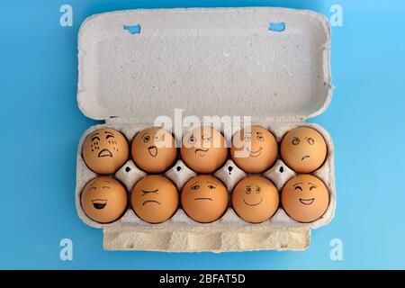 Brown eggs in a cardboard tray top view. Eggs with the image of different emotions on a blue background. Chicken eggs with painted faces. Stock Photo