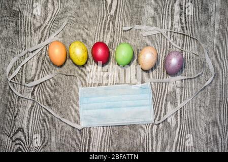 Many colored easter eggs laying near medical mask on the wooden background. Coronavirus Covid-19. Concept photo. Easter 2020. Stock Photo