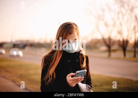 Young woman wearing protective face medical mask while using smart phone walking on street in city