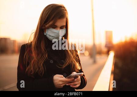Young woman wearing protective face medical mask using smart phone while standing on bridge in city at sunset Stock Photo