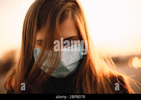 Close up portrait of sad depressed young woman wearing protective face medical mask while standing in city during sunset Stock Photo