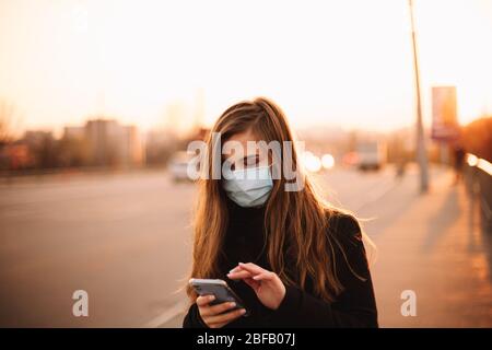 Happy young woman wearing protective face medical mask using smart phone while walking on empty sidewalk on bridge in city at sunset Stock Photo