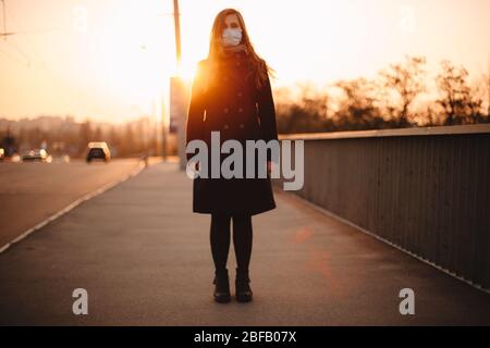 Portrait of young woman wearing protective face medical mask while standing on empty sidewalk on bridge in city during sunset Stock Photo