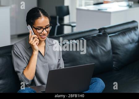 Beautiful woman using a smartphone having phone call from her apartment. Businesswoman enjoying conversation sitting on a couch. Stock Photo