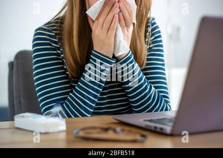 Sick woman working on laptop at home, covering nose and mouth when coughing and sneezing in paper tissue, ill freelance woman with cold working