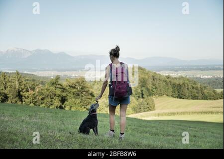 View from behind of a young female hiker, standing in green meadow with her black dog next to her, enjoying the view. Stock Photo
