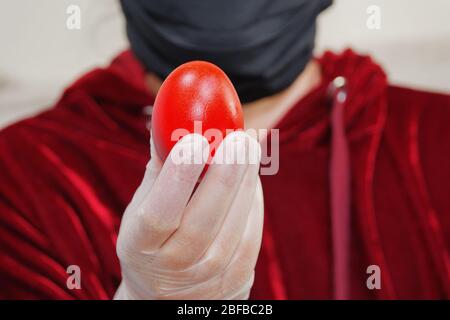Person holding red Easter hard-boiled egg before camera. Unidentified female wearing black face mask and white protective gloves displays painted egg. Stock Photo