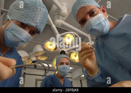 A surgical team of three doctors – two men and one woman – wearing blue gowns, caps and masks, inside a hospital operating theatre underneath overhead Stock Photo
