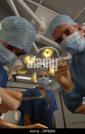 A surgical team of three doctors – two men and one woman – wearing blue gowns, caps and masks, inside a hospital operating theatre underneath overhead Stock Photo