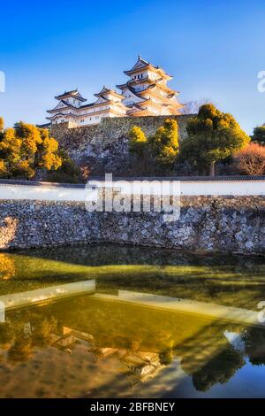 Water moat around stone walls of main white tower in Himeji on a sunny day against blue sky. Stock Photo