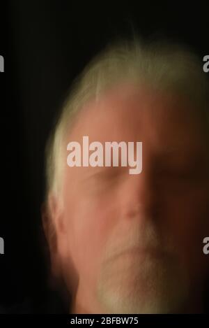 The blurred face of an older man with white hair and a goatee with eyes closed in a meditative state against a black background. Stock Photo
