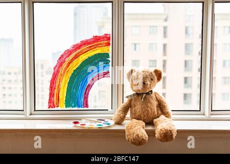 Toy and painted rainbow during Covid-19 quarantine at home. Girl near window. Stay at home Social media campaign for coronavirus prevention, let's all Stock Photo