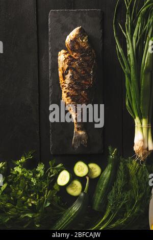 Dorado fish cooked with cucumbers, fresh onions, parcel and dill on a black wooden surface Stock Photo