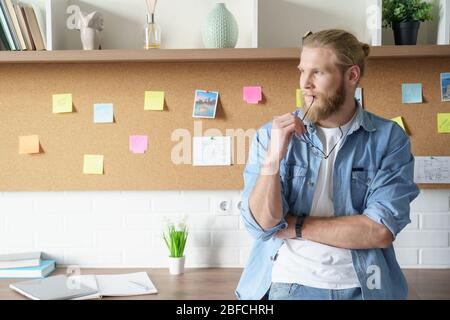 Young man freelancer looking away dreaming standing at home office. Stock Photo