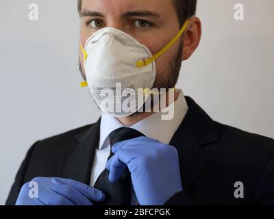 A businessman wearing an N95 respirator dust mask adjusts a black tie while wearing blue nitrile rubber gloves and a suit in a closeup view. Stock Photo