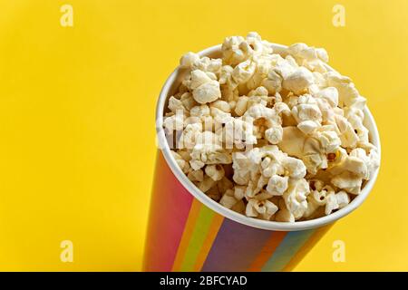 Popcorn in papercup. Multi coloured striped paper cup bucket with popcorn against a yellow background with copy space. Cinema or movies concept. Stock Photo