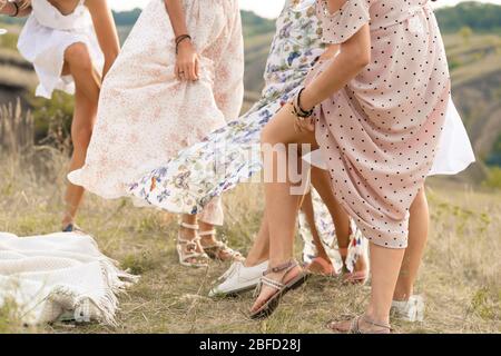 The company of female friends having fun, dancing on summerfield in long dresses showing their legs. Summer rural style picnic concept