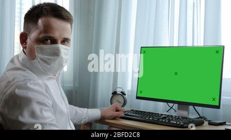 Medium shot. View to the display. Business man in medical mask looking seriously to camera sitting next to the blank display. Green Screen Mock-up Dis Stock Photo