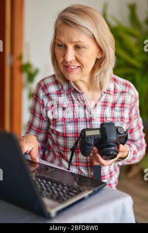 Portrait of attractive mature female photographer holding camera, looking at laptop Stock Photo