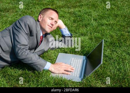 Lazy businessman relaxing on a sunny patch of green summer grass falling asleep at his laptop computer Stock Photo