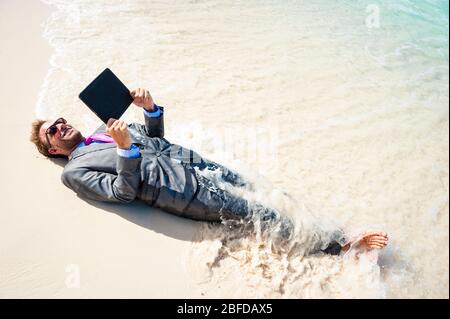 Barefoot businessman relaxing with a tablet computer on the shore of a tropical beach with waves breaking over his suit Stock Photo