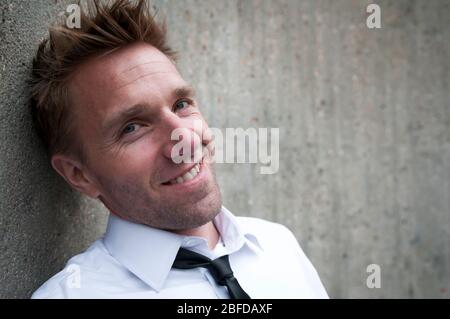 Portrait of man in smart casual outfit of white dress shirt and skinny back tie smiling outdoors in front of a textured concrete wall Stock Photo