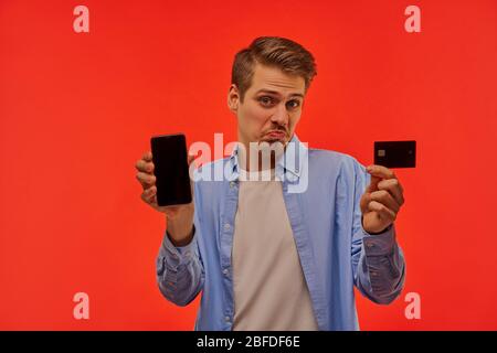Disappointed guy with a beard shows a smartphone and a credit card. Stock Photo