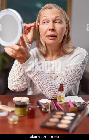 pretty, aged woman applying black mascara, dye eyelashes using tassel, making, doing professional make Stock Photo