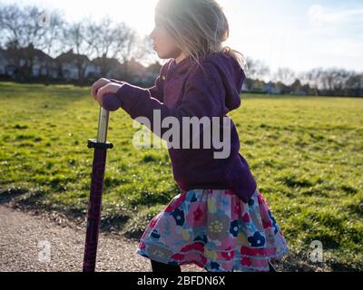 Four year old girl moving fast on scooter through a sunny London park in Spring. Stock Photo