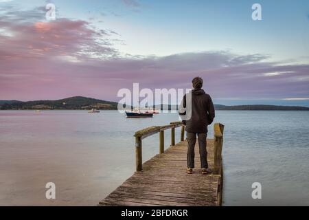 Tourist standing on an old wooden jetty viewing the picture perfect sunset on the harbour, with boats safely anchored in St Helen''s Bay in Tasmania. Stock Photo