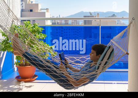 Boy with digital tablet computer lying in the hammock hanging on balcony. Education, online distance learning, homeschooling for kids Stock Photo