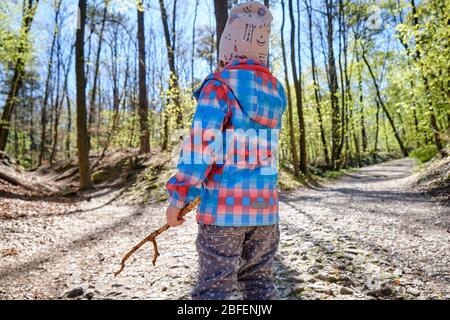 Rear view of 4 year old child girl holding wooden stick in hand and thinking about which way to choose in a beautiful bright springtime forest on a su Stock Photo
