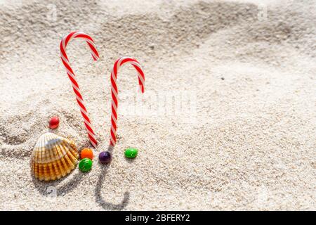 candy cane, seashells and colorful candies on white sand background. copy space. Stock Photo
